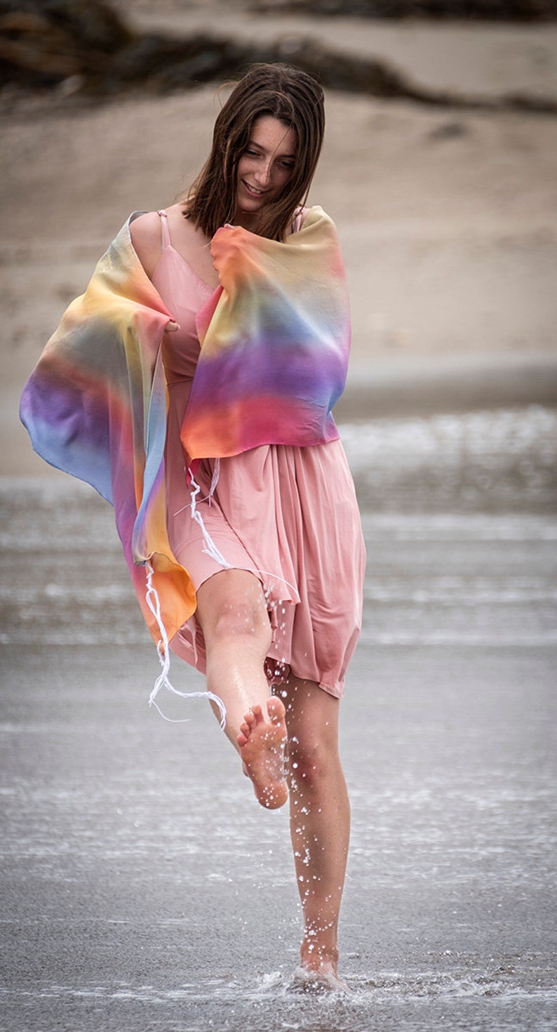 May include: A woman wearing a pink dress and a colorful shawl stands in shallow water on a beach. She is smiling and kicking her foot in the water, creating a splash.