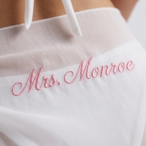 May include: White sheer fabric with the embroidered text "Mrs. Monroe" in a delicate pink script. The fabric is gathered at the top with white ties. The overall aesthetic is soft and feminine.