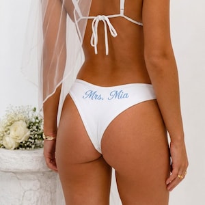 Puede incluir: Braguita de bikini blanca con el texto "Mrs. Mia" en escritura azul claro. La braguita forma parte de un conjunto nupcial, con un top y un velo a juego visibles. La imagen está tomada desde atrás, mostrando la parte trasera de la braguita.