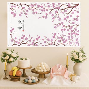 May include: A banner with Korean text and cherry blossom branches, set above a table with a cake, fruit, and traditional Korean clothing. The scene includes white roses, candles, and a variety of foods, creating a festive atmosphere.