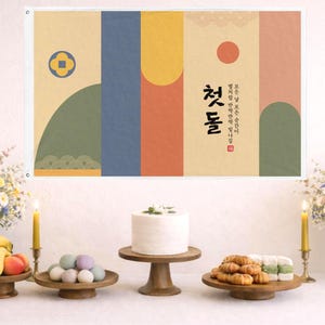 May include: A festive table setting with a banner featuring Korean text and abstract shapes in muted colors. The table is adorned with flowers, candles, a cake, and various treats, creating a celebratory atmosphere.