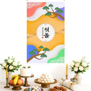 May include: A colorful banner with Korean text and a decorative design featuring pine branches and abstract shapes. The banner hangs above a table set with traditional Korean food, cakes, and flowers, creating a festive atmosphere.