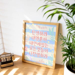 Puede incluir: Una impresión enmarcada con texto coreano en un marco de madera. La impresión tiene un fondo azul claro con rayas blancas y detalles florales. A la izquierda se ve un pequeño calendario de escritorio.
