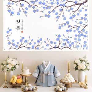 May include: A Korean traditional first birthday celebration scene. A banner with blue floral branches and Korean text hangs above a table. The table displays a baby's light blue hanbok, a small cake, fruit, and traditional rice cakes.