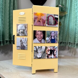 May include: A yellow miniature locker with a black handle and several square magnets attached. The magnets display various photographs, including images of a dog, children, and flowers. The locker is on a light-colored wooden surface.