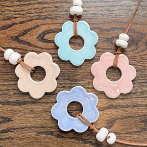Könnte beinhalten: Vier Keramik-Blumenanhänger in Pastellfarben: Hellblau, Rosa, Lavendel und Beige. Jeder Anhänger hat ein zentrales Loch und ist an einer braunen Lederschnur mit weißen Perlen befestigt. Präsentiert auf einer Holzoberfläche.