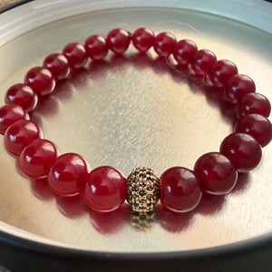 May include: A red beaded bracelet with a gold-colored, textured bead in the center. The bracelet is made of round, polished red beads. The bracelet is displayed on a silver-colored surface.