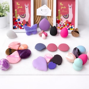 May include: An assortment of makeup sponges and accessories in various shapes and colors, including teardrop, heart, and butterfly designs. The image also features framed artwork with a llama illustration and the text "No prob llama."