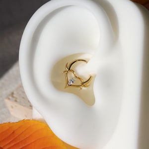 May include: A gold-colored, heart-shaped earring with a small, clear gemstone in the center. The earring has a unique design with curved accents. The earring is shown on a white ear model.