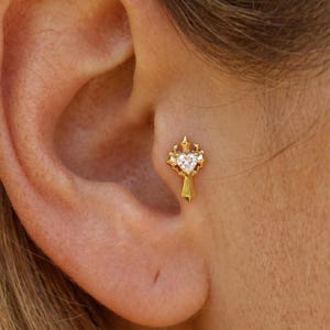 May include: A gold-toned ear stud featuring a central heart-shaped clear gemstone set within a decorative, pointed cross-like frame. The jewelry piece is positioned in the tragus area of the ear, showcasing a polished metallic finish and intricate detailing.