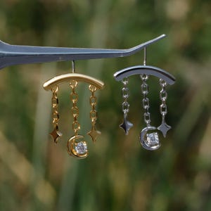 Puede incluir: Dos pendientes de tem&aacute;tica celestial, uno dorado y otro plateado, est&aacute;n suspendidos. Cada pendiente presenta una barra curva, colgantes de cadena, dijes de estrella y una luna creciente que sostiene una gema transparente. El fondo es verde suave.