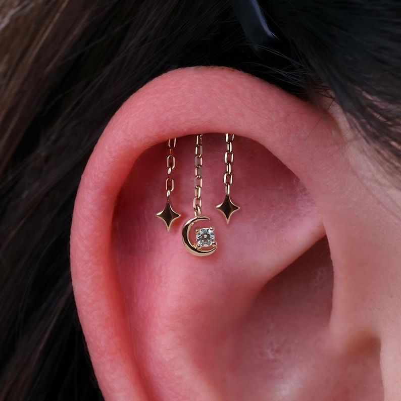 Puede incluir: Joya de piercing de oreja dorada con una luna creciente con una gema central y tres cadenas colgantes. Cada cadena termina con un dije en forma de estrella. La joya se usa en la oreja.