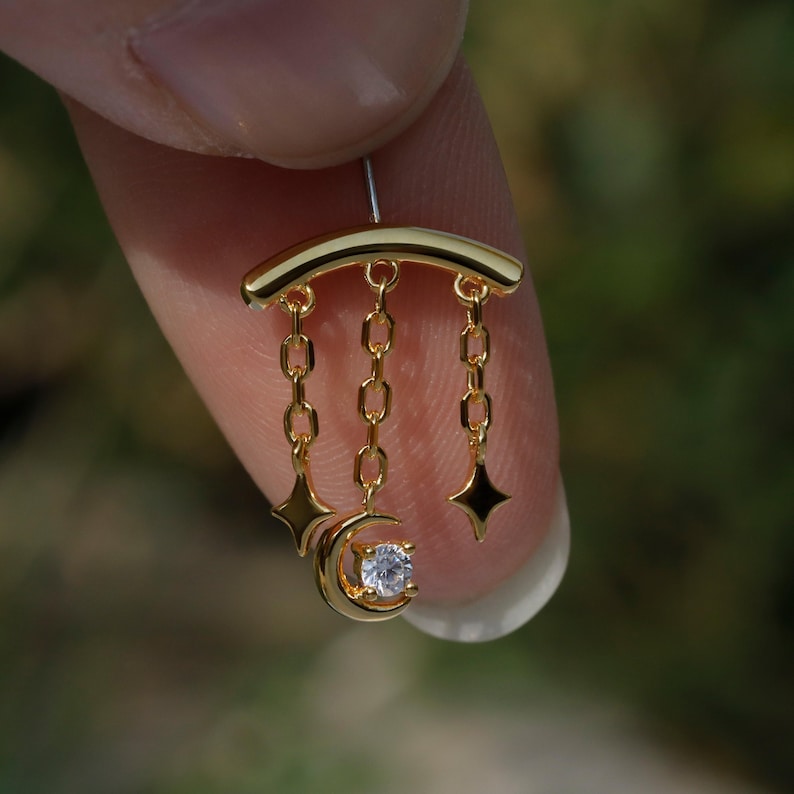 Puede incluir: Un pendiente dorado con un dise&ntilde;o celestial. Tiene una barra curva en la parte superior, con tres cadenas colgando. Cada cadena tiene una estrella o una luna con una gema transparente. El pendiente est&aacute; sujeto por un dedo.