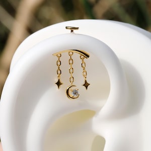 Pode incluir: Joia de orelha dourada com uma barra curva, correntes penduradas, estrelas e uma lua crescente com uma gema central. A joia é colocada sobre um fundo branco, destacando seu design celestial.