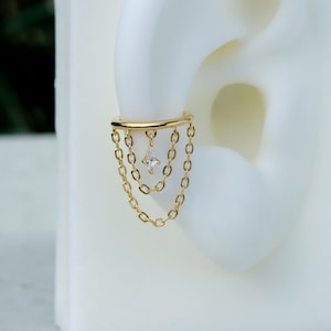 May include: A gold-colored ear cuff featuring a dangling chain design. The cuff has a curved bar that attaches to the ear, with three chains hanging down. A diamond-shaped, clear gemstone is suspended from the center chain.