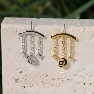 Puede incluir: Un par de pendientes de tem&aacute;tica celestial. Uno es plateado con un dise&ntilde;o de cadena, una luna con una piedra transparente y detalles de estrellas. El otro es dorado con un dise&ntilde;o similar, con colgantes de luna y estrella.