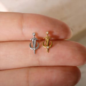 Könnte beinhalten: Zwei kleine Ohrringe, einer silberfarben und einer goldfarben, jeweils mit einem Schwertdesign, einem Halbmond und einem kleinen Diamanten. Die Ohrringe werden vor einem hautfarbenen Hintergrund gezeigt.