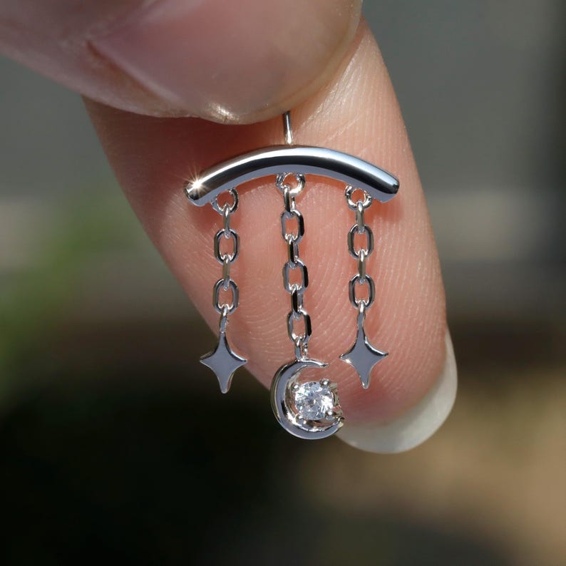 Puede incluir: Un colgante plateado con una barra curva y tres cadenas. Cada cadena tiene un dije: dos dijes en forma de estrella y una luna creciente con una gema clara en el centro. El colgante est&aacute; sujeto por un dedo.