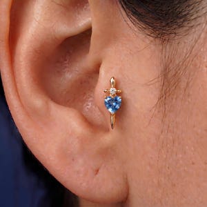 May include: A gold-coloured earring shaped like a dagger, featuring a light blue heart-shaped gemstone. A small, clear gemstone is set above the heart. The earring is worn on the ear.