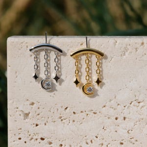 Puede incluir: Pendientes plateados y dorados con tem&aacute;tica celestial. Cada pendiente presenta una barra curva, colgantes de cadena, dijes de estrella y una luna creciente con una gema central. Se muestran sobre una superficie texturizada y neutra.