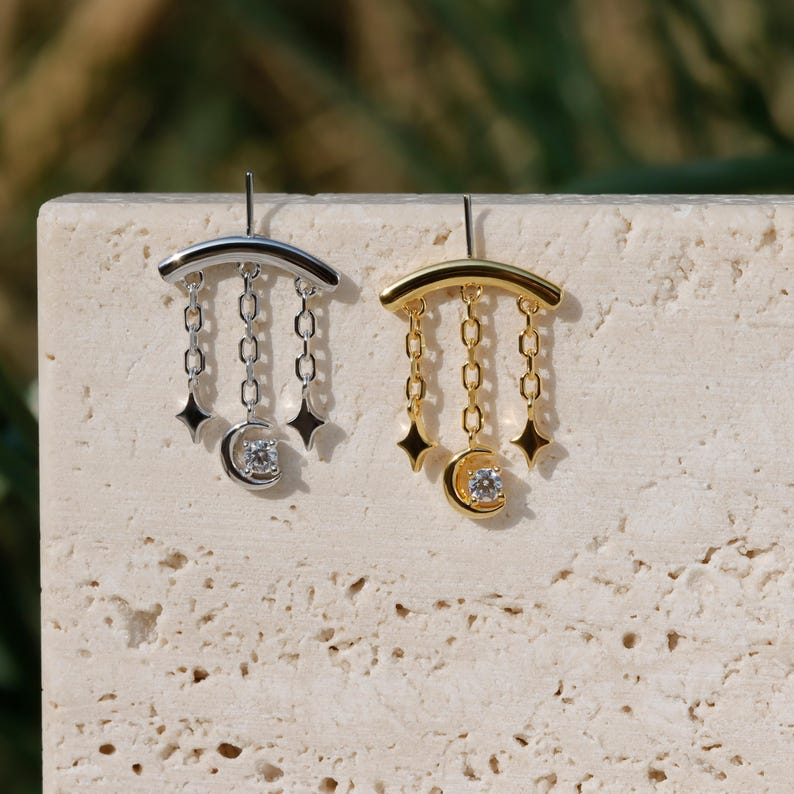 Puede incluir: Pendientes plateados y dorados con tem&aacute;tica celestial. Cada pendiente presenta una barra curva, colgantes de cadena, dijes de estrella y una luna creciente con una gema central. Se muestran sobre una superficie texturizada y neutra.