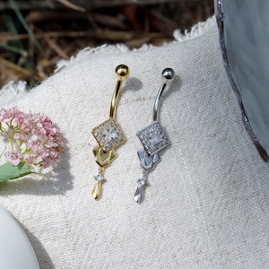 Może przedstawiać: Dwa kolczyki do pępka. Jeden jest w kolorze złotym z kwadratowym, diamentowym środkiem i wiszącym urokiem w kształcie łzy. Drugi jest w kolorze srebrnym o podobnym wzorze. Oba są prezentowane na białej, teksturowanej powierzchni.