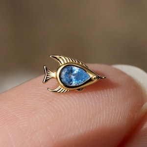 Puede incluir: Un pendiente en forma de pez de tono dorado con una piedra preciosa azul claro en forma de pera en el centro. El cuerpo metálico del pez muestra detalles de líneas grabadas en las aletas y la cola, con un pequeño punto circular como ojo.
