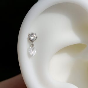 May include: A silver earring with a clear, round gemstone at the top and a dangling heart-shaped gemstone. The earring is shown against a white background, highlighting its delicate design and sparkle.