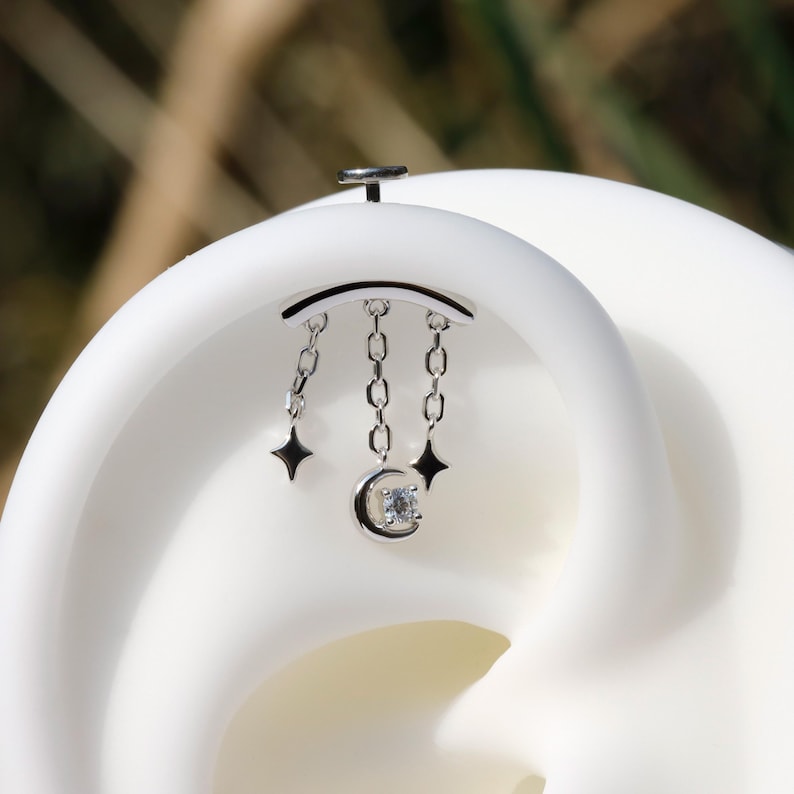 Puede incluir: Un piercing de oreja plateado con una barra curva y cadenas colgantes. Cada cadena tiene una estrella o una luna creciente con una peque&ntilde;a gema transparente. La joya est&aacute; sobre un fondo blanco.