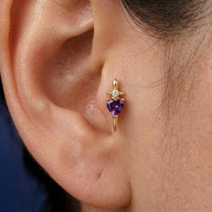 Könnte beinhalten: Ein goldfarbener Ohrring in Form eines Dolches, mit einem herzförmigen lila Edelstein und einem kleinen Diamanten. Der Ohrring wird im Ohr getragen und zeigt sein detailliertes Design und seine funkelnden Elemente.