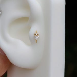 Peut inclure: Une boucle d'oreille dorée avec une pierre précieuse en forme de cœur et une petite pierre précieuse ronde au-dessus. La boucle d'oreille a la forme d'un poignard. La boucle d'oreille est présentée sur un modèle d'oreille blanc.