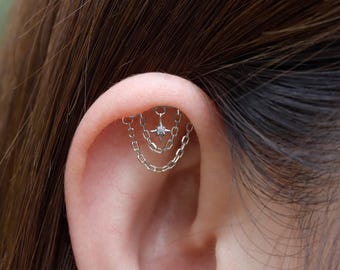 Peut inclure: Un piercing d'oreille argenté avec un charm en forme d'étoile orné d'une petite pierre précieuse transparente, suspendu par de fines chaînes. Les chaînes pendent d'une barre incurvée, créant un effet superposé. Le bijou est placé sur l'oreille d'une personne.