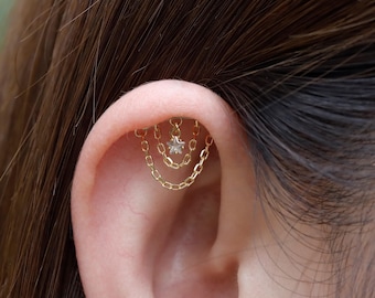 Peut inclure: Un piercing d'oreille doré avec trois chaînes pendantes. La chaîne supérieure porte une petite gemme claire en forme d'étoile. Le piercing est porté sur le cartilage supérieur de l'oreille.