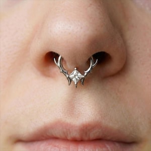 May include: A silver septum ring with a deer antler design and a central square, clear gemstone. The nose ring is shown in situ, highlighting the detailed metalwork and the sparkling gemstone. The design is intricate and eye-catching.
