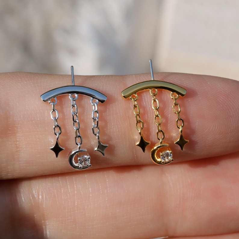 Puede incluir: Dos pendientes de tem&aacute;tica celestial, uno plateado y otro dorado. Cada pendiente presenta una barra curva con cadenas colgantes, estrellas y una luna creciente con una peque&ntilde;a gema transparente. Dise&ntilde;ados para orejas perforadas.