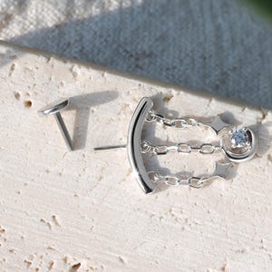 Puede incluir: Un par de pendientes plateados. Un pendiente presenta un dise&ntilde;o de barra curva con tres eslabones de cadena, una estrella y una peque&ntilde;a gema redonda y transparente. El otro pendiente es un peque&ntilde;o y plano perno ovalado.