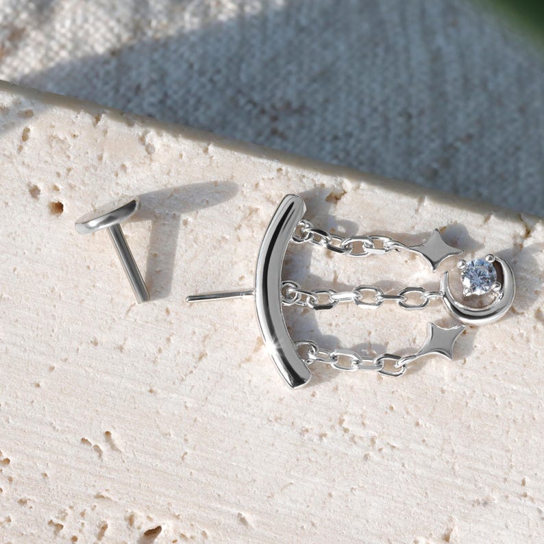 Puede incluir: Un par de pendientes plateados. Un pendiente presenta un dise&ntilde;o de barra curva con tres eslabones de cadena, una estrella y una peque&ntilde;a gema redonda y transparente. El otro pendiente es un peque&ntilde;o y plano perno ovalado.