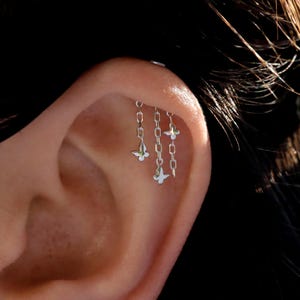 May include: A silver-toned ear cartilage piercing featuring three delicate dangling chains of varying lengths. Each chain is adorned with a small, polished butterfly charm at the end, creating a layered, cascading effect along the outer ear rim.