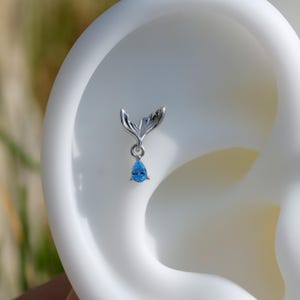 May include: A silver earring with a mermaid tail motif and a blue, teardrop-shaped gemstone pendant. The earring is displayed against a white backdrop, emphasizing the delicate design and the gemstone's colour.