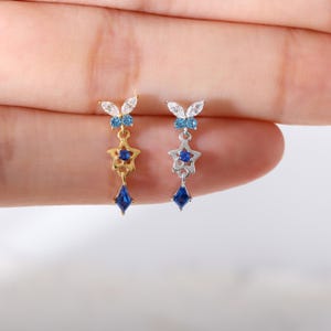 May include: Two dangle earrings, one gold-toned and one silver-toned, each featuring a butterfly top with white and light blue stones, a central star-shaped charm with a blue stone, and a bottom kite-shaped blue stone drop pendant.
