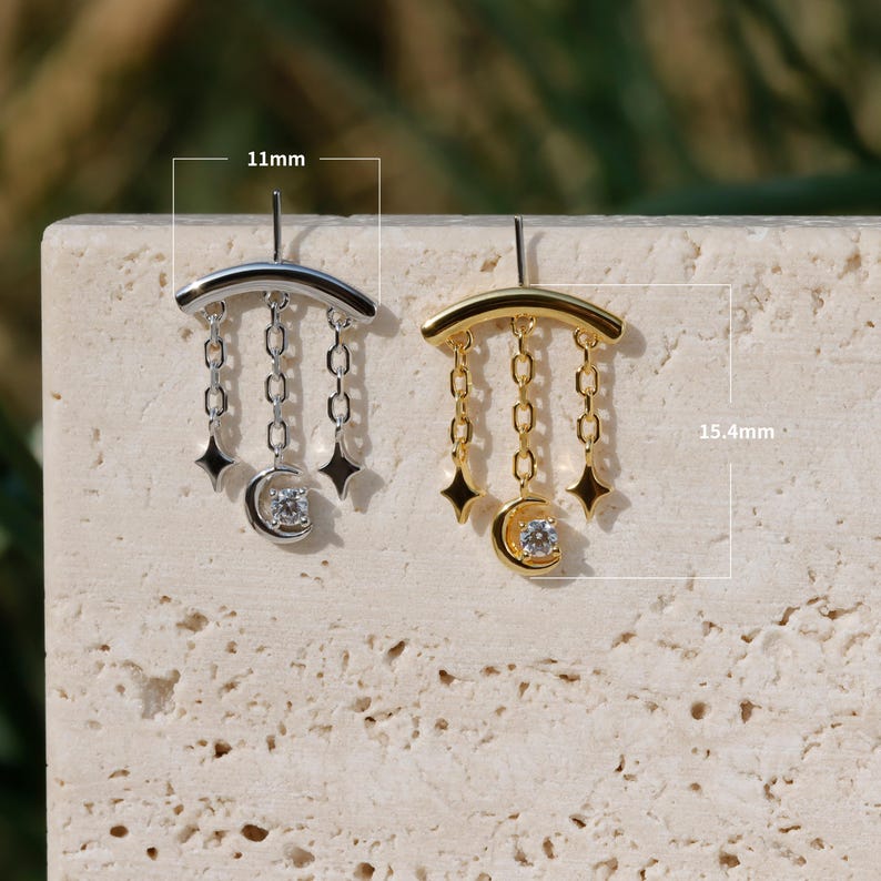 Puede incluir: Pendientes plateados y dorados. Cada pendiente presenta una barra curva, colgantes de cadena, abalorios de estrella y una luna creciente con una gema transparente. Los pendientes miden 1,1 cm de ancho y 1,54 cm de largo.