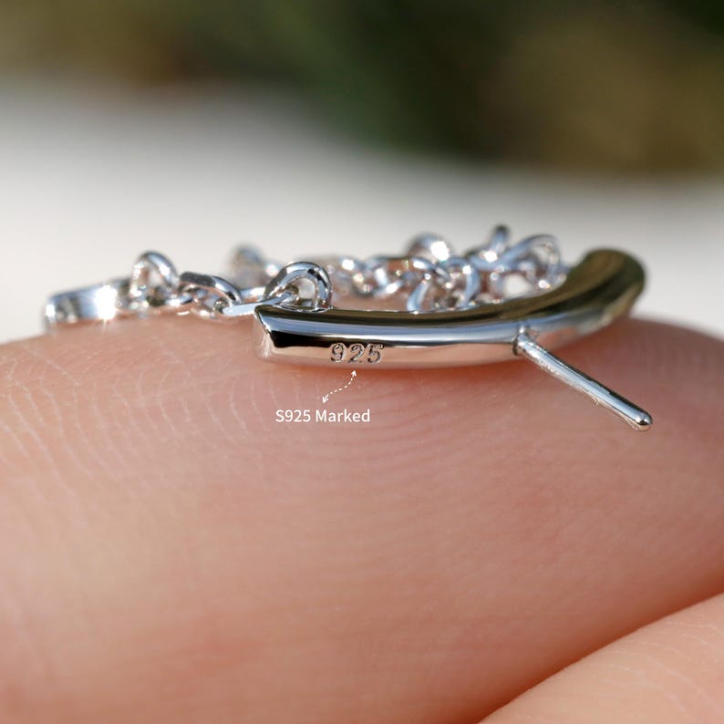 Puede incluir: Primer plano de un pendiente plateado con un dise&ntilde;o curvo y un poste. El pendiente est&aacute; marcado con "925" y el texto "S925 Marked". El pendiente est&aacute; en un dedo.