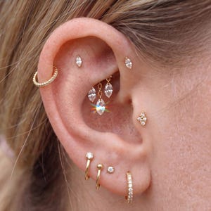 Könnte beinhalten: Eine Sammlung goldfarbener Ohrpiercings mit einem texturierten Ring, mehreren Ohrsteckern mit klaren, marquise-geschliffenen Edelsteinen, einem Cluster-Stecker und hängenden Elementen, verteilt auf Helix, Conch und Ohrläppchen.