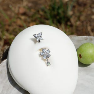 May include: Two silver belly button rings with clear crystal accents. One ring features a butterfly design, while the other has a flower with a dangling teardrop gem. The jewelry is displayed on a white surface.