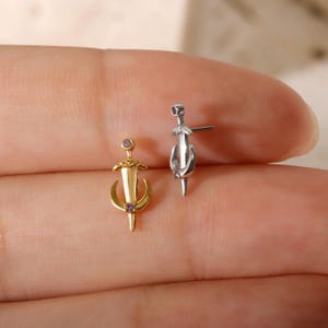Op de afbeelding: Twee oorknopjes, één goudkleurig en één zilverkleurig, elk met een zwaardontwerp met een paarse edelsteen accent. De gouden oorbel heeft een halve maan vorm. De oorbellen worden gepresenteerd op een huidskleurige achtergrond.