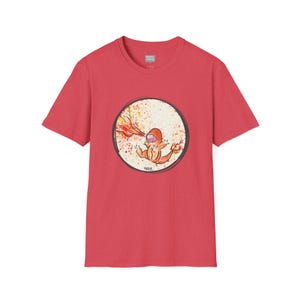 May include: Red t-shirt featuring a circular graphic of a cartoon character with orange and yellow flames. The character is inside a white circle with a black border and paint splatter effect. The shirt is made of a soft material.
