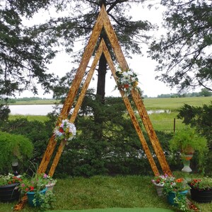 Könnte beinhalten: Ein hölzerner Hochzeitsbogen in Dreiecksform, verziert mit Blumenarrangements. Der Bogen ist im Freien auf einem grünen Rasen aufgestellt, mit Topfpflanzen und einem See im Hintergrund. Der Bogen ist aus hellbraunem Holz.