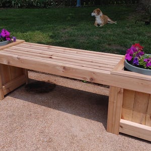 Peut inclure: Banc en bois avec deux jardinières intégrées remplies de fleurs colorées. Le banc est en bois clair et possède une assise à lattes. Un chien est couché sur l'herbe en arrière-plan.