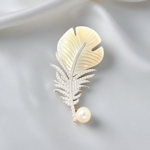 May include: A feather-shaped brooch with a pearl accent. The brooch features a large, cream-colored feather design with intricate detailing. The stem and smaller feather details are encrusted with sparkling crystals, and a single pearl hangs at the bottom.
