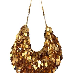 Puede incluir: Bolso de hombro dorado con lentejuelas, de forma curva. El bolso está cubierto de pequeñas lentejuelas redondas doradas. Tiene una fina correa marrón y un interior blanco.